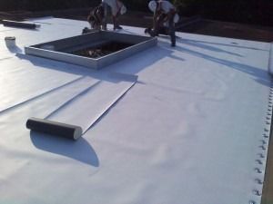 Two workers installing white roofing material on a flat roof around a skylight.