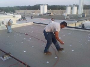 Two workers install white circular fasteners on a flat grey commercial roof under a clear sky.