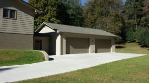 A tan house with a two-car garage, a concrete driveway, and a lawn, set against a backdrop of trees.
