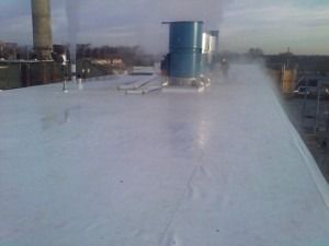A flat commercial roof with a white membrane surface, featuring blue exhaust vents and a tall chimney under a cloudy sky.