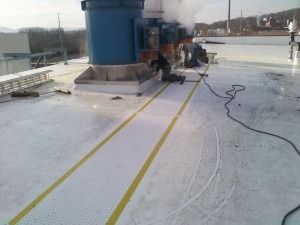 A worker performs maintenance on large blue industrial rooftop equipment next to a yellow-bordered walkway.