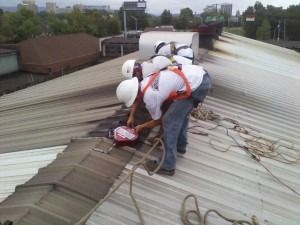 Two workers in white helmets and safety harnesses working on a sloped metal roof, securing a red fall-protection device.