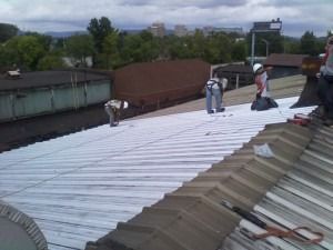 Workers in hard hats and safety gear install a white protective layer onto a sloped metal roof outdoors.