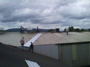 Workers repair a metal roof on a building under a cloudy sky.
