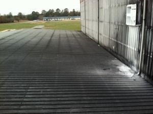 A ribbed metal roof surface extends toward a grassy field and trees, adjacent to a textured exterior wall.