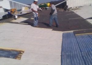 Two construction workers install black roofing material on a white flat roof during the day.