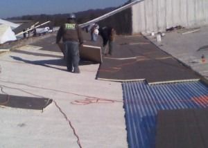 Two workers install insulation panels on a large, flat construction site roof featuring corrugated metal sections.