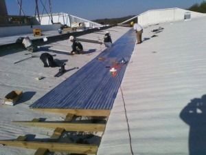 Construction workers install blue corrugated metal roof panels on a sloped white commercial roof structure.