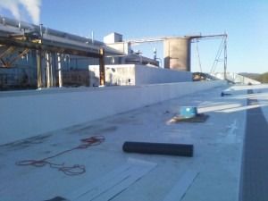 A flat, light-colored roof under construction with industrial pipes, silos, and steam vents against a clear blue sky.
