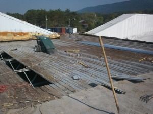 Construction scene on a flat roof with metal decking, a cutting machine, and white angled roof sections against a hillside.
