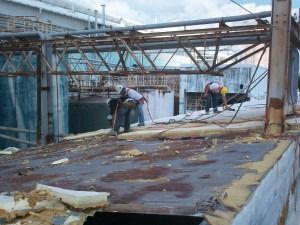 Two workers in hard hats and harnesses repair a rusted industrial roof structure.