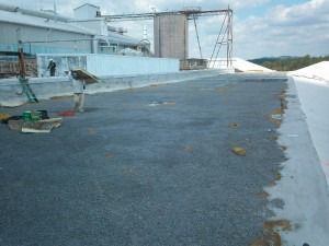 A worker walks across a large, dark-surfaced industrial roof near a tall storage structure under a partly cloudy sky.