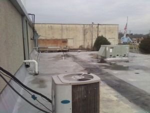 Two HVAC units sit on a gray commercial rooftop near a low wall, with a building and flag visible in the background.
