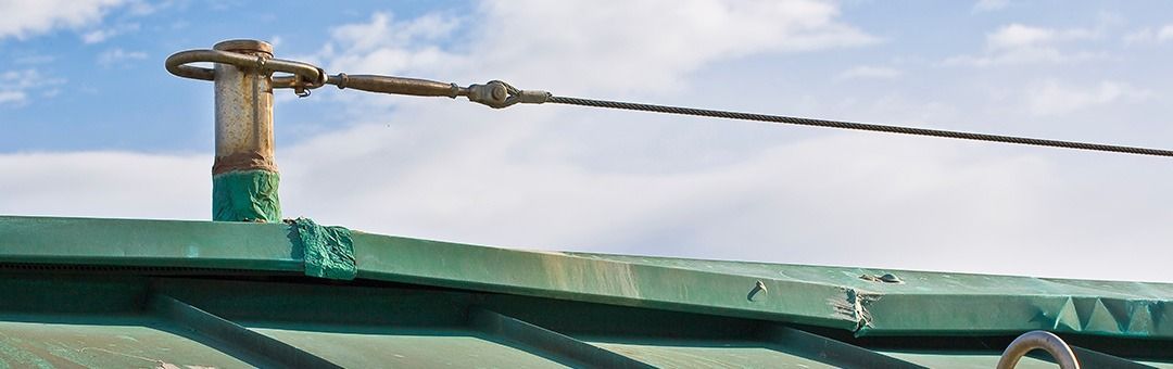 A metal post secured to a green roof with a thin, twisted cable attached to it against a cloudy blue sky.