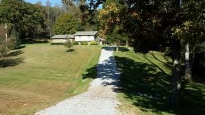 A gravel driveway leads through a grassy yard toward a two-story light-colored house surrounded by mature trees.
