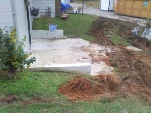 A view of a house exterior showing a concrete patio, a small retaining wall, and disturbed soil from construction work.