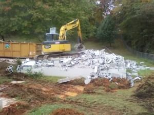 A yellow excavator works at a demolition site, clearing a pile of white concrete rubble near a large yellow dumpster.