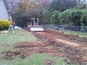 A bulldozer levels ground in a residential yard near a survey tripod, with a house and trees in the background.
