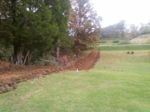 A freshly excavated dirt path runs along the edge of a grassy lawn bordered by trees on an overcast day.