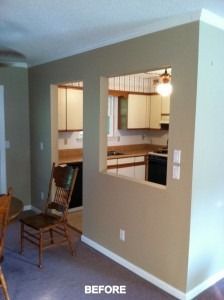 A tan wall with two large pass-through openings revealing a kitchen with white cabinets and a dark wooden chair nearby.