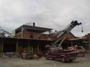 A construction crane operates at a brick industrial building with a red pickup truck and a forklift parked in front.