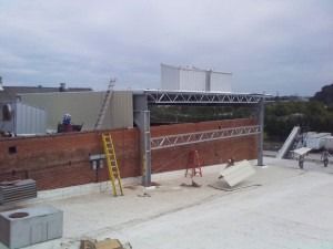 Construction workers assemble a metal framework on the flat roof of a red brick building under an overcast sky.