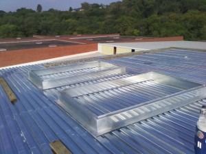 Two rectangular metal skylight frames installed on a blue corrugated metal roof against a backdrop of trees.