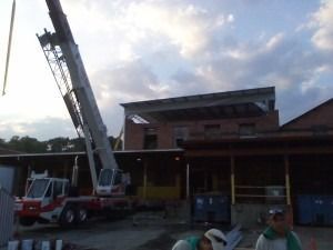 A crane lifts construction materials toward the roof of a two-story brick building at dusk.