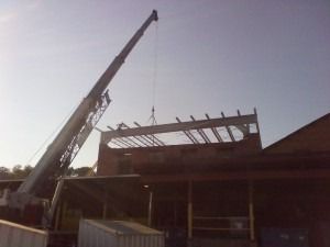 A construction crane lifts a large steel beam onto the roof of a brick building.