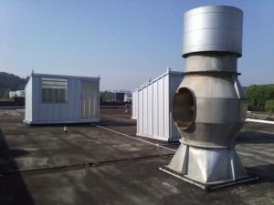 A rooftop with two small sheds and a large, cylindrical industrial exhaust fan under a clear blue sky.