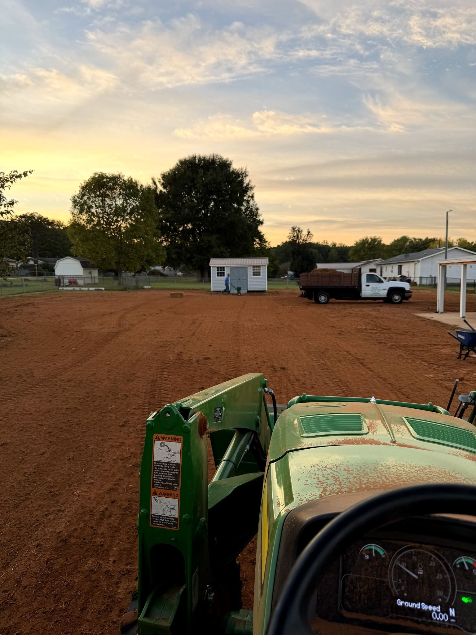 Green tractor in a gravel lot at sunset, with small trailers, trees, and a white truck in the distance.