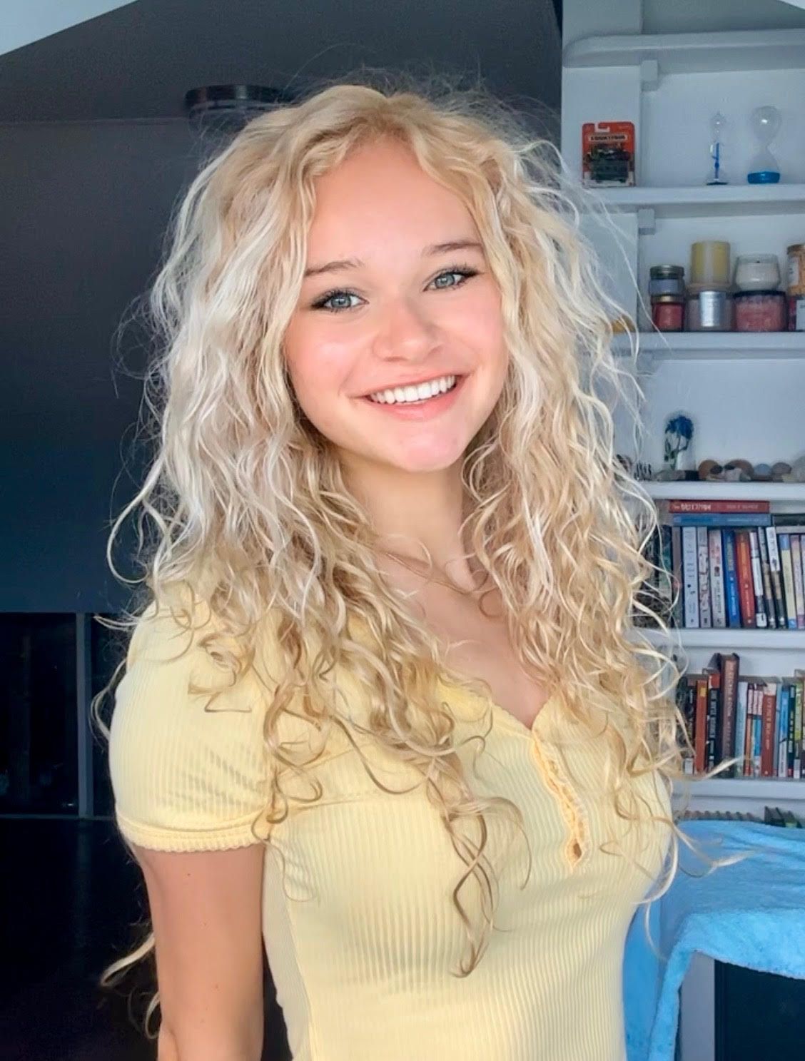 Blonde woman with curly hair smiling, wearing a yellow top. Standing near a shelf with books.