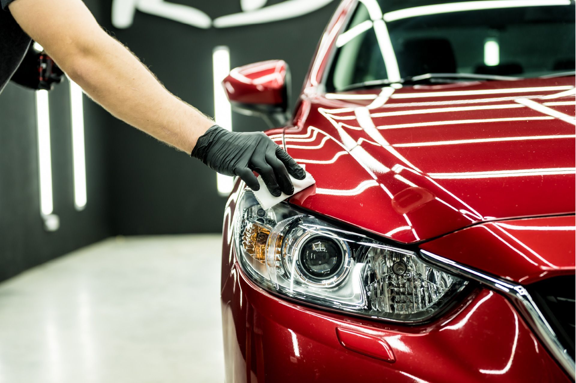 Person in black gloves applying polish to a red car headlight in a detailing shop.