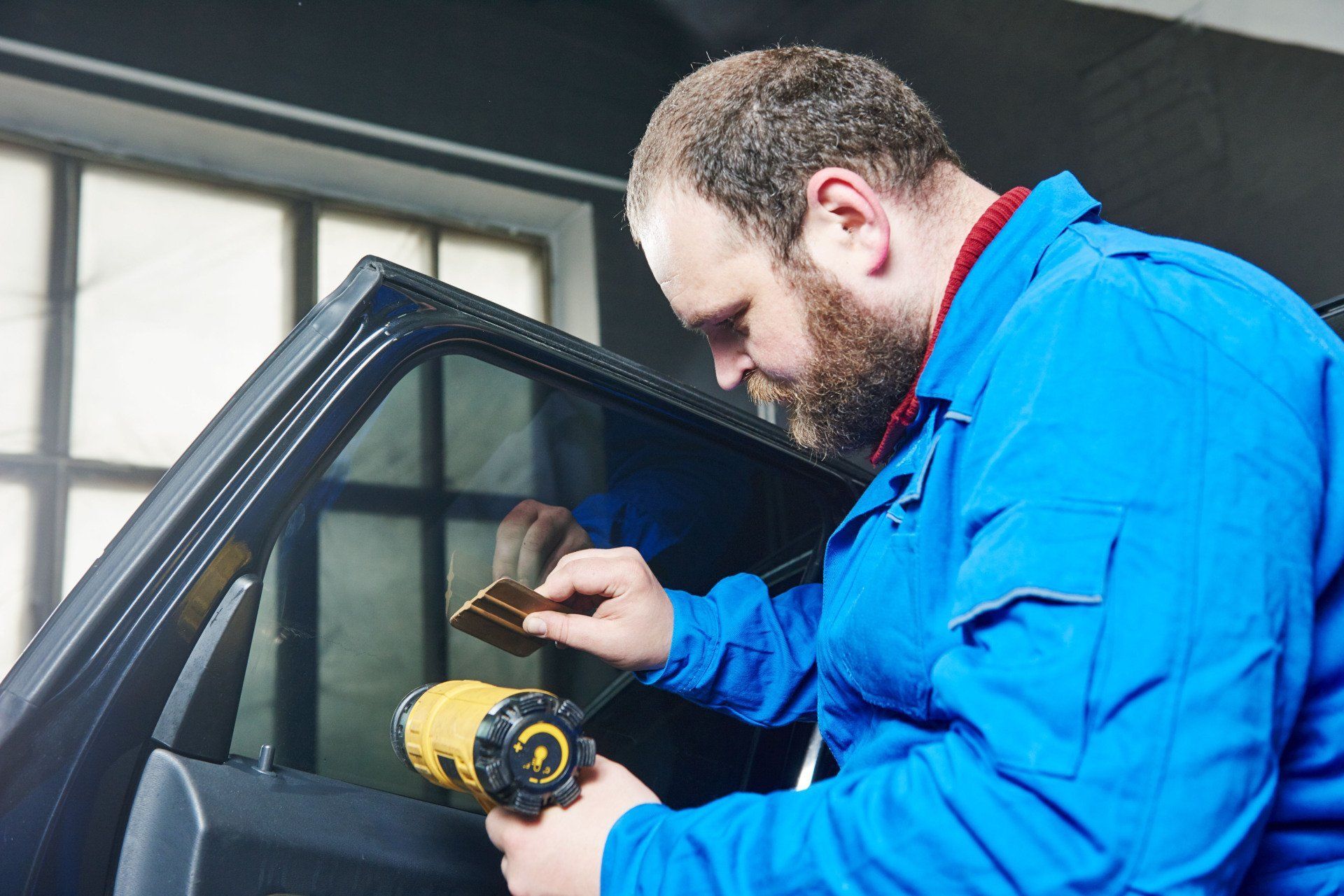 Man in blue jumpsuit applying tint to a car window with a tool and heat gun in a garage.