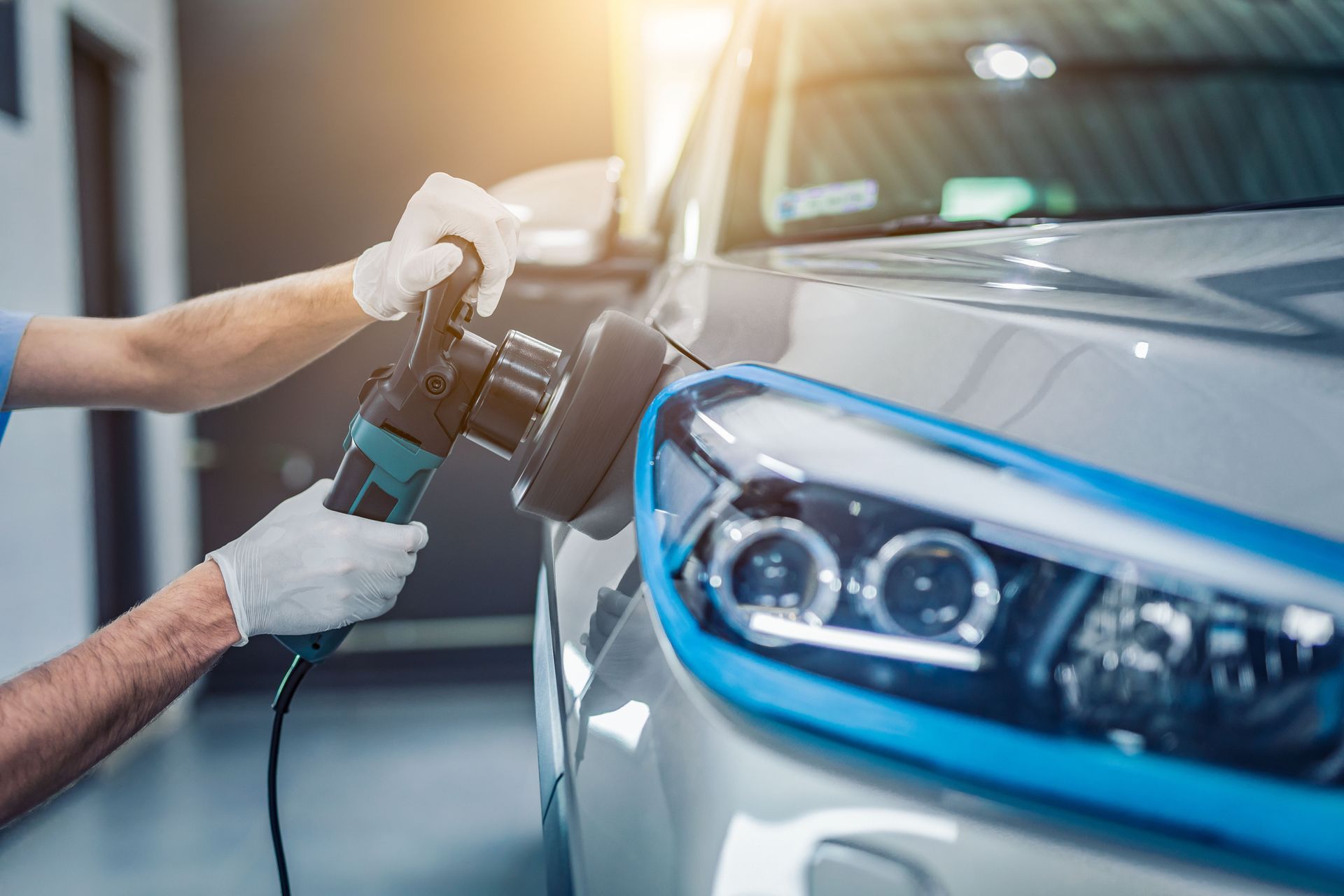 A person polishes a silver car with a power buffer in a garage.