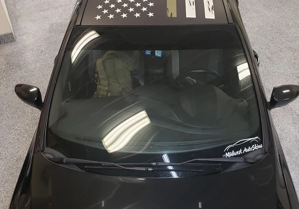 Black car with a windshield banner and a thin blue line American flag decal on the roof.