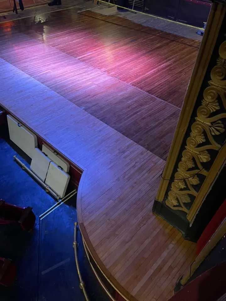 Wooden stage with curved edge, bathed in purple and red light; empty theater seating visible below.