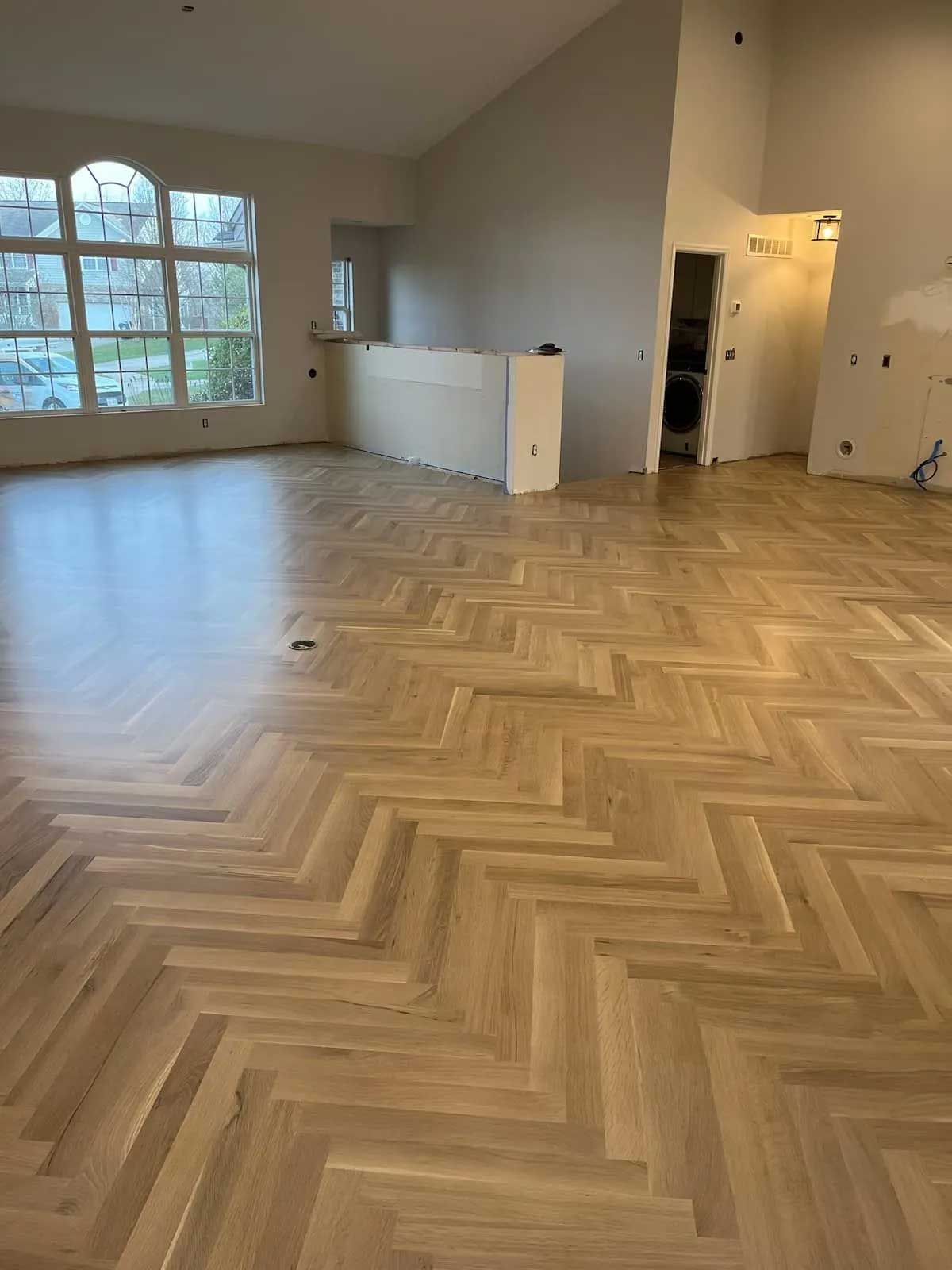 Herringbone pattern wood floor in an empty room with a large window and white walls.