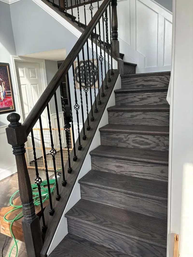 Wooden staircase with dark gray treads and a black wrought-iron banister. White wall paneling.