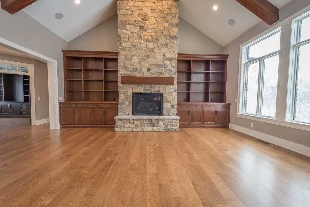 Empty living room with wood floors, stone fireplace, built-in shelves, and large windows.