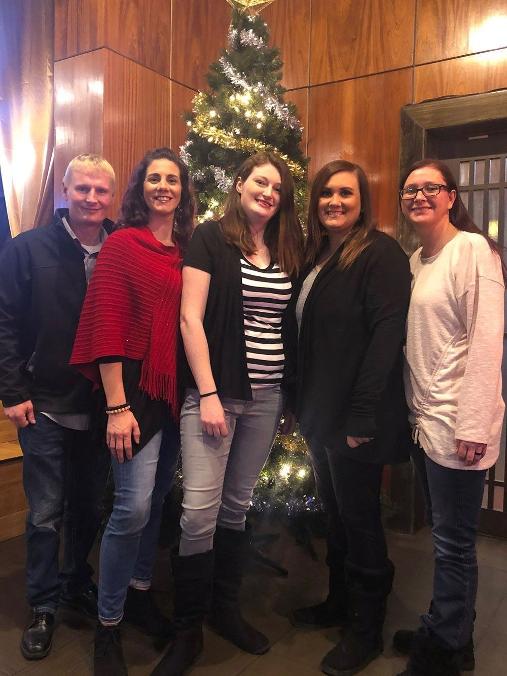 Five people posing indoors beside a lit Christmas tree in a wood-paneled room