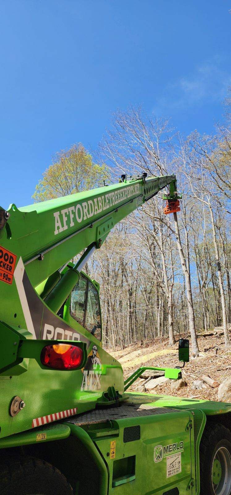 A green crane is lifting a tree in the woods.