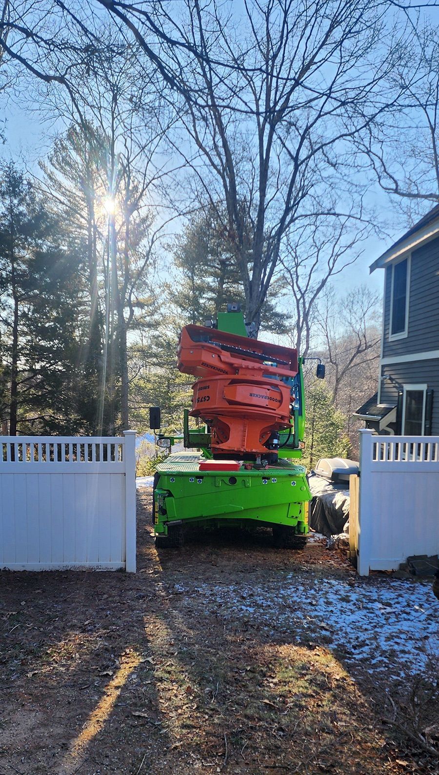 A green and orange machine is cutting a tree in front of a house.