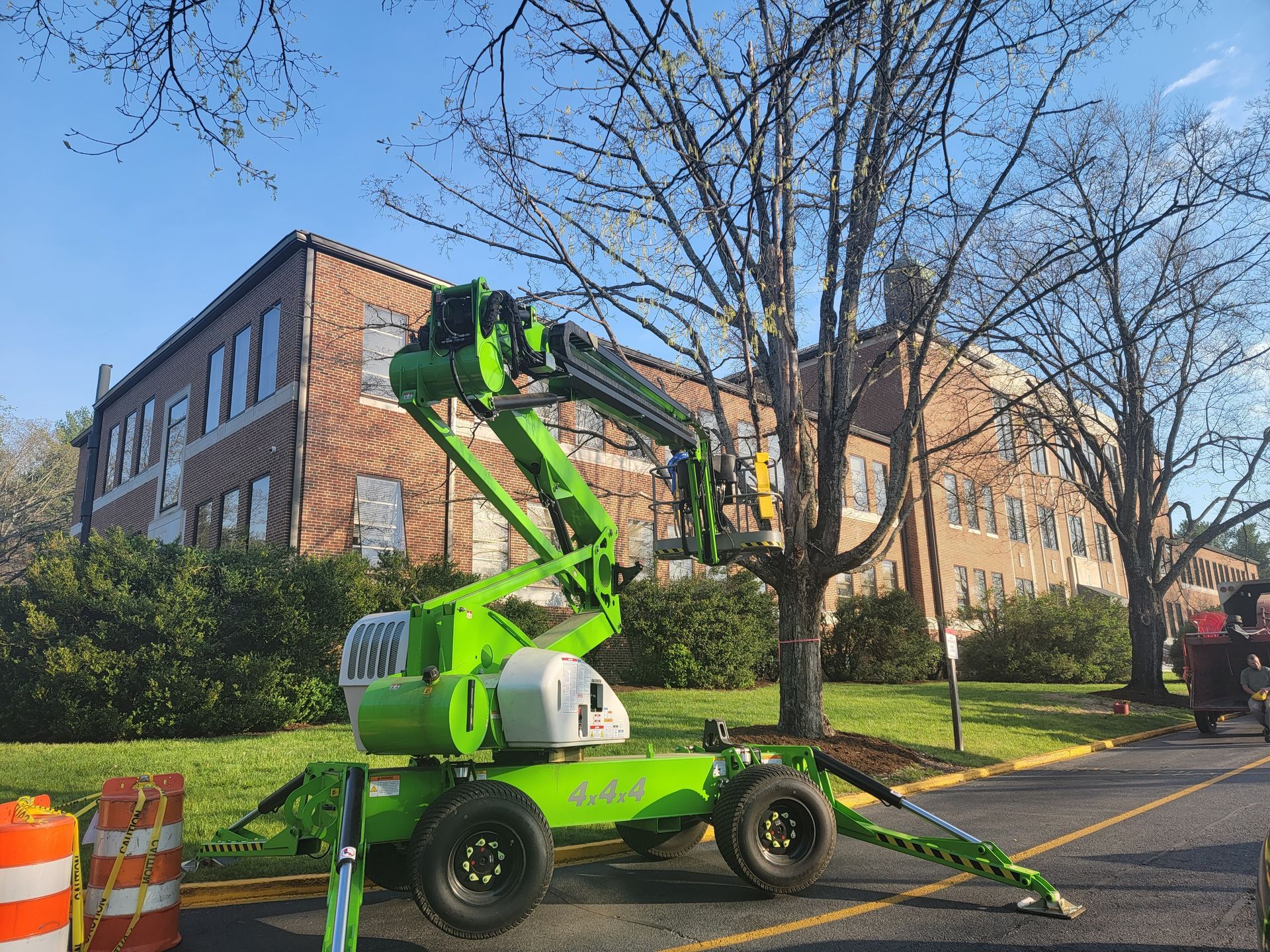 Green aerial lift near a brick building and a tree, parked on a paved surface.
