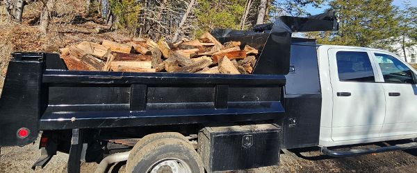 A black dump truck loaded with split firewood, parked on a dirt road.