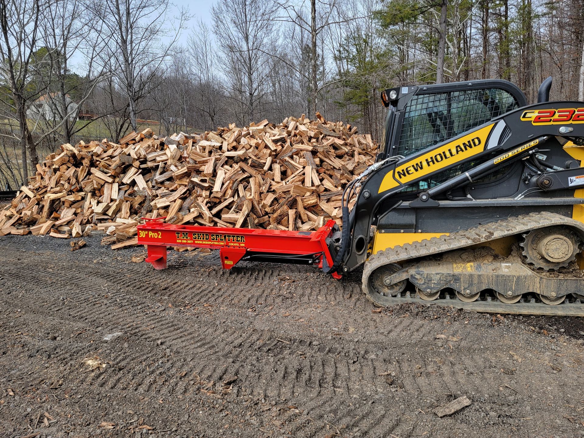 Yellow New Holland skid steer with a red log splitter, wood pile in background.