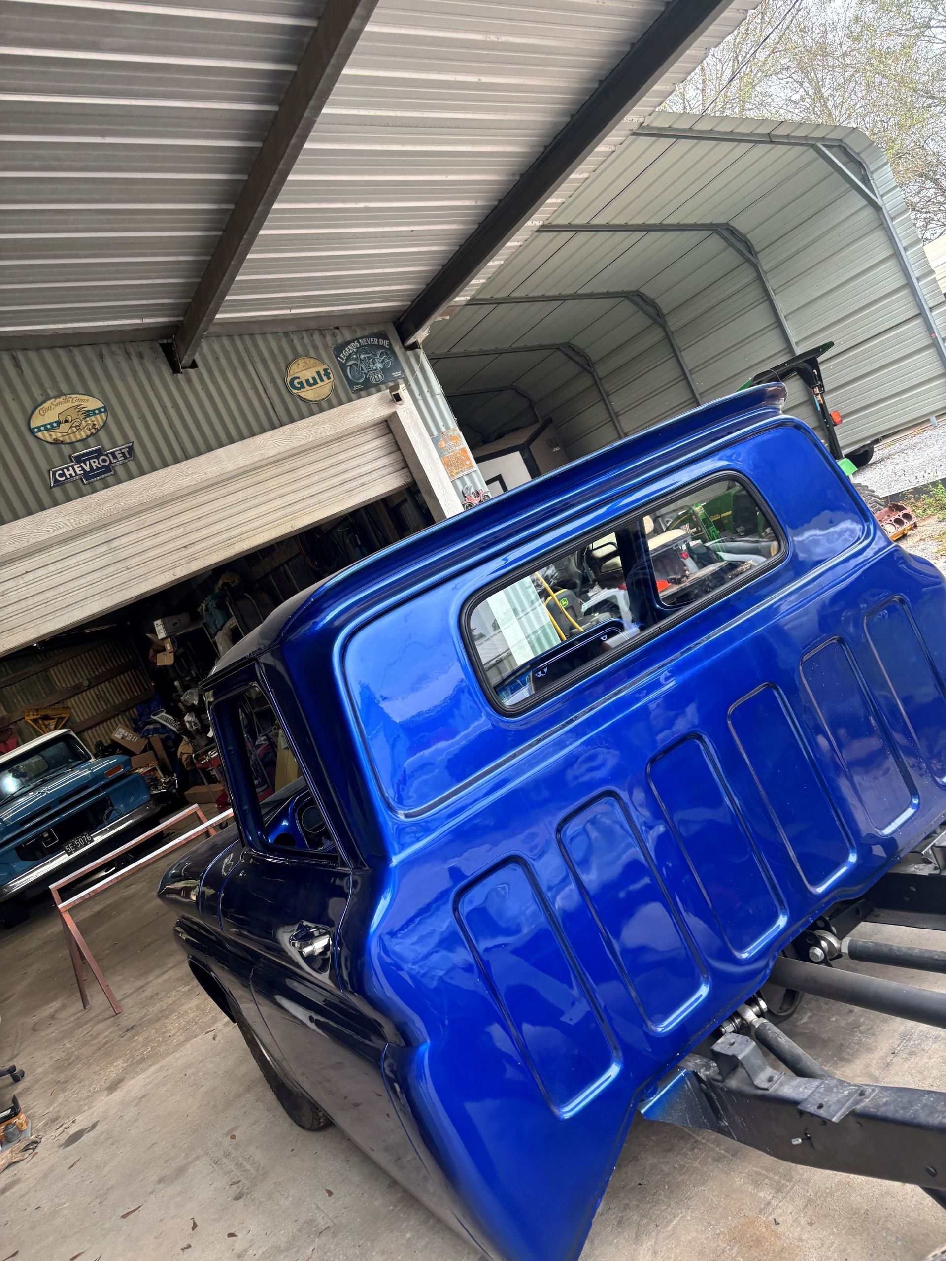 A bright blue truck cab sits on a frame under a metal workshop awning.