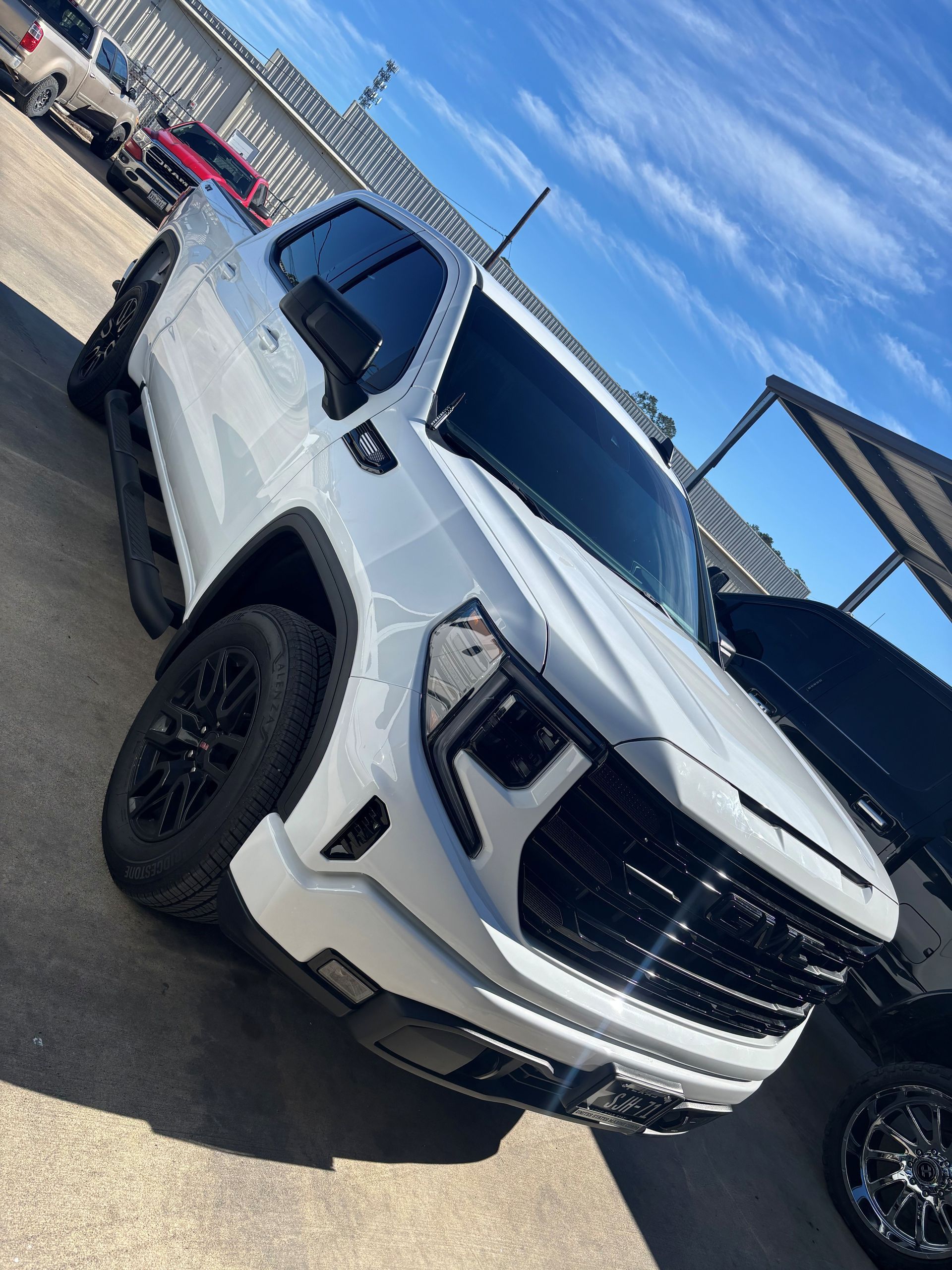 A white GMC Sierra pickup truck with black trim and wheels parked on an asphalt lot under a blue sky.