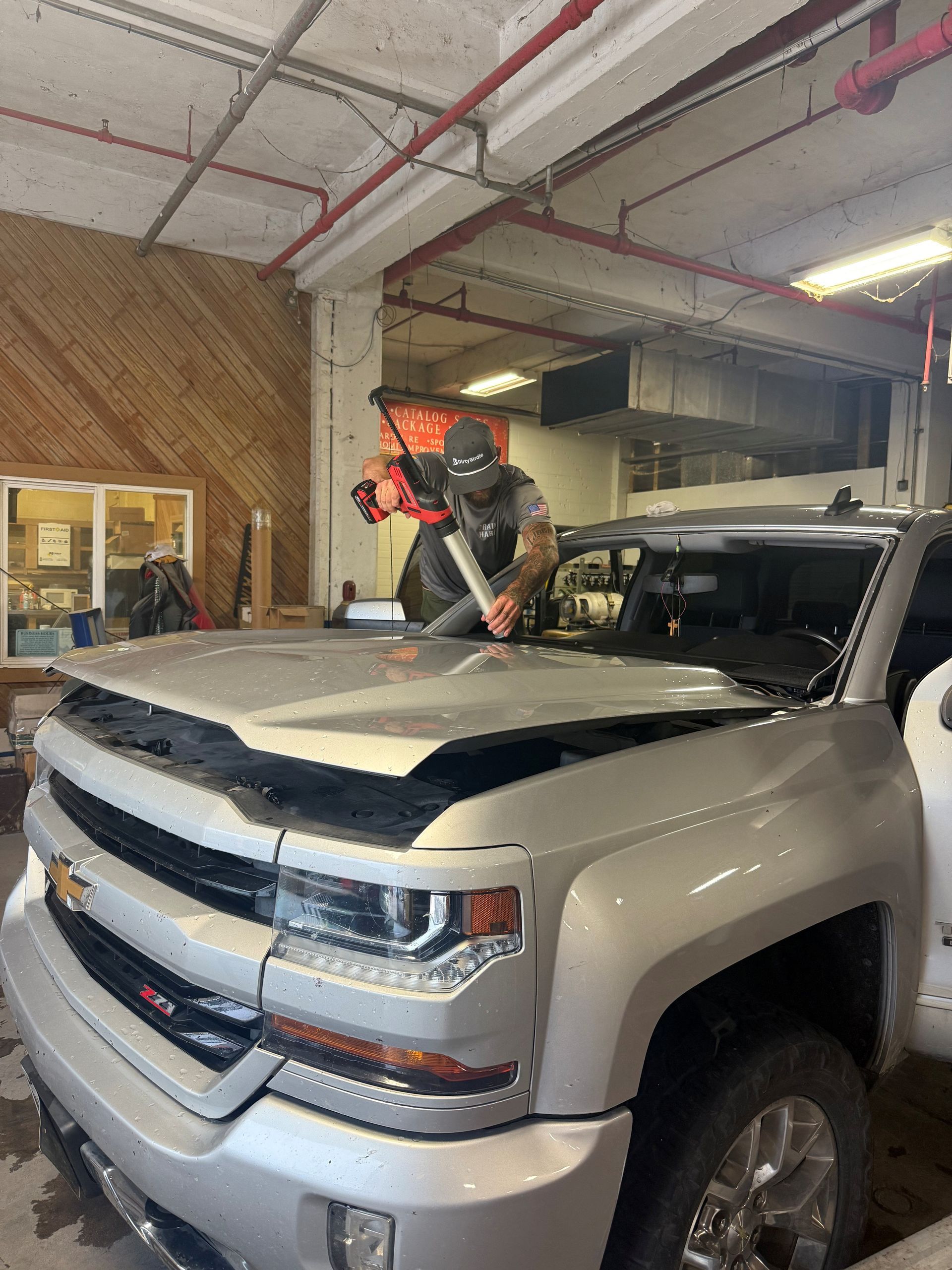 A person in a garage uses a power tool to work on the windshield of a silver pickup truck with the hood open.