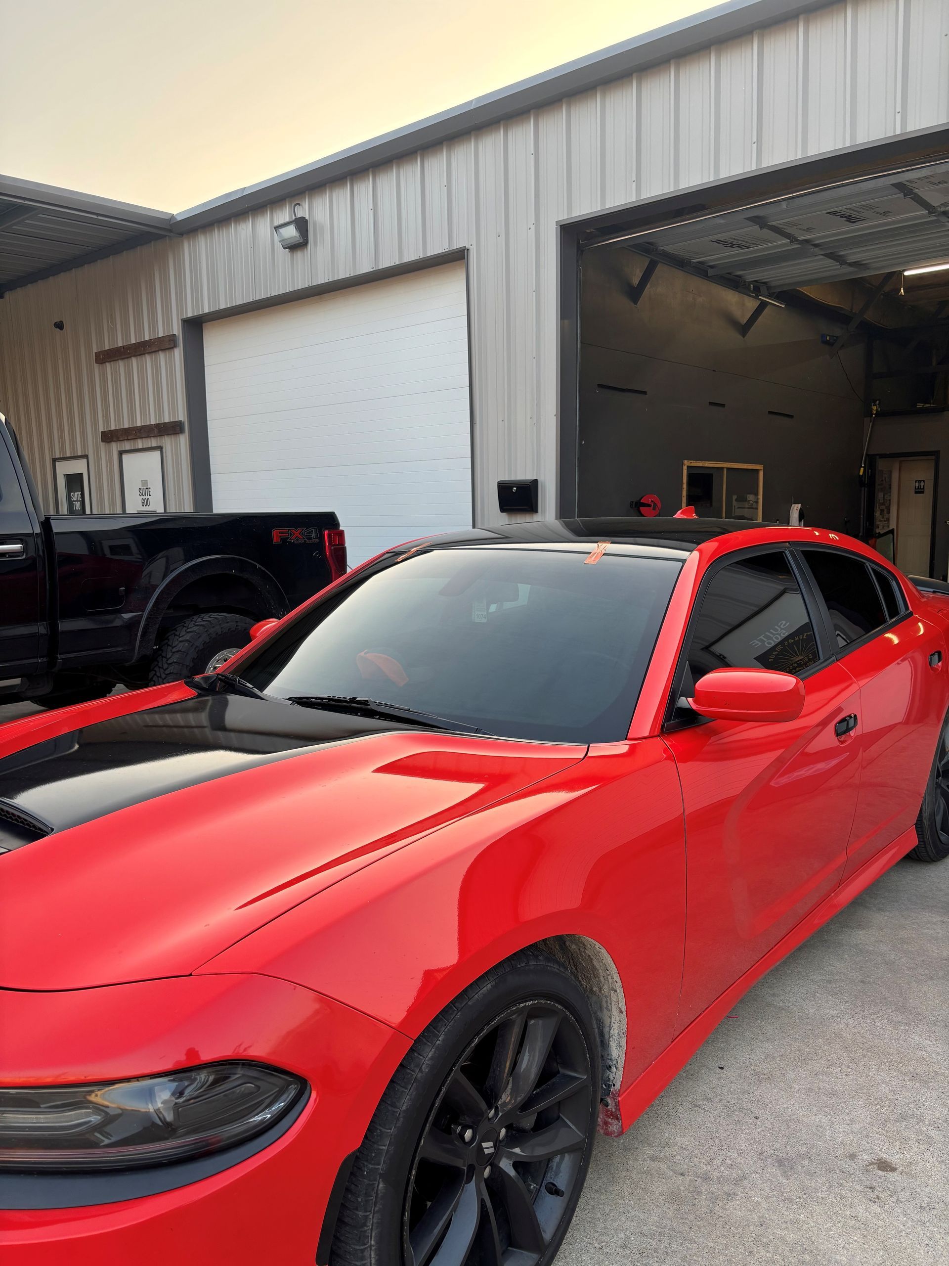 A red Dodge Charger with a black roof and hood stripe parked outside a garage next to a black truck.